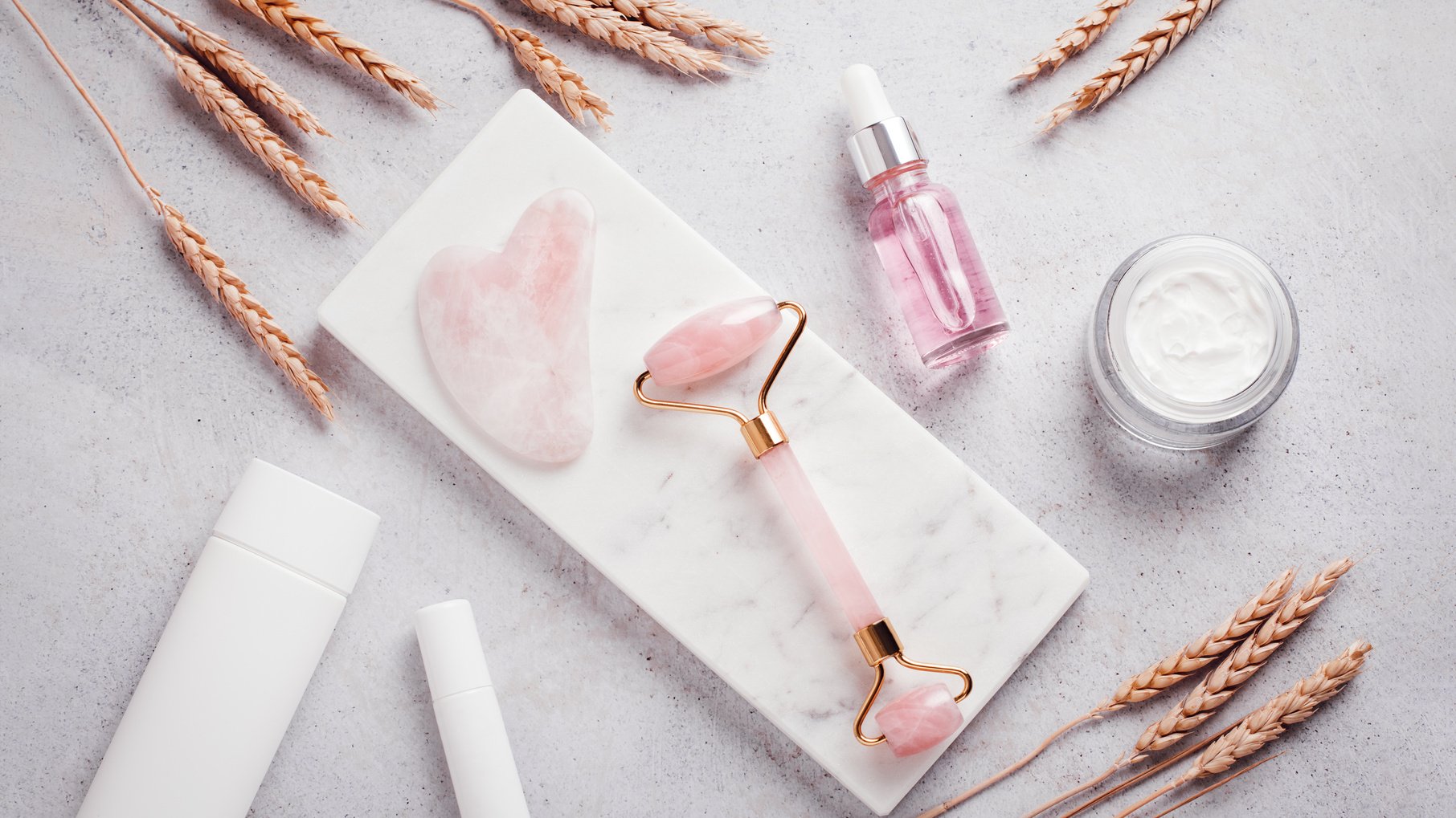 Facial roller, gua sha, serum and moisturizing cream on marble background